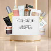 Cohorted Beauty Box mystery box with multiple skincare and makeup products arranged behind it on a beige pedestal in a warm indoor setting. Gifts for her, UK, beauty box, cosmetics, makeup, skincare, ESPA, Grow Gorgeous, Avant, OFRA