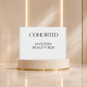 Cohorted Beauty Box mystery box with multiple skincare and makeup products arranged behind it on a beige pedestal in a warm indoor setting. Gifts for her, UK, beauty box, cosmetics, makeup, skincare, ESPA, Grow Gorgeous, Avant, OFRA